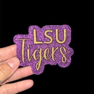 May include: A purple glittery patch with the words "LSU Tigers" in gold script. The patch has a unique shape and is held against a black background. The text is embroidered.
