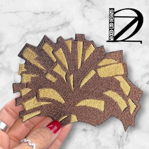 May include: A brown and gold embroidered patch with a stylized design of a tree with a sunburst pattern. The patch is made of a soft, textured fabric.