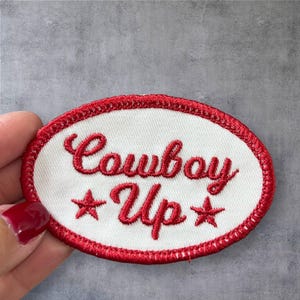 May include: White oval embroidered patch with red stitching. The patch says "Cowboy Up" in red cursive lettering with a red star on either side of the word "Up".