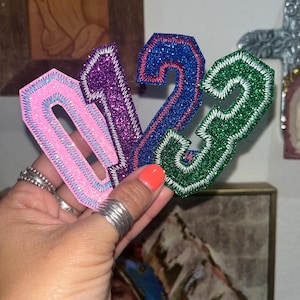 May include: A set of four glittery iron-on patches featuring the numbers zero, one, two, and three. The zero is light pink, the one is purple, the two is blue, and the three is green. Each number has a thick, stitched border in a contrasting thread color.