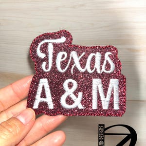 May include: A red glitter iron-on patch with white embroidery that reads "Texas A&M".