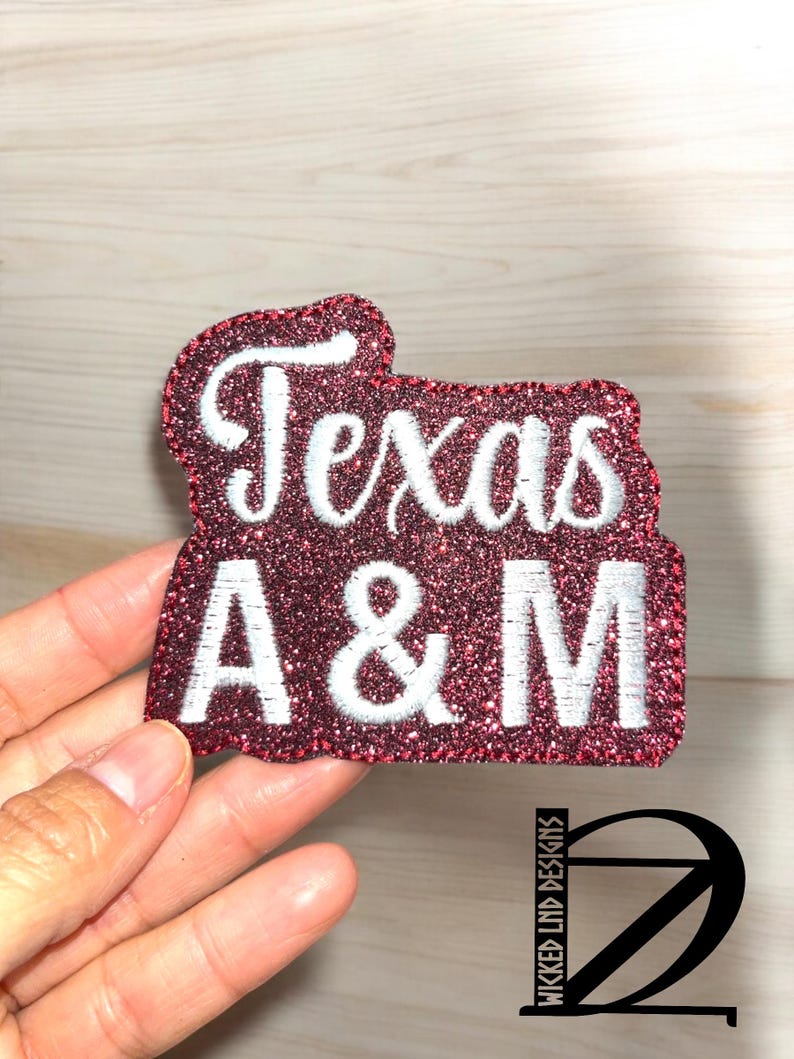 May include: A maroon glitter fabric patch featuring the white embroidered text Texas A&M. The patch has a die-cut shape that follows the contour of the lettering, with a dense, sparkling maroon surface and clean white stitching for the letters.