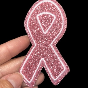 May include: A pink glitter ribbon patch with a white outline, held against a black background. The ribbon is a symbol of awareness and support.