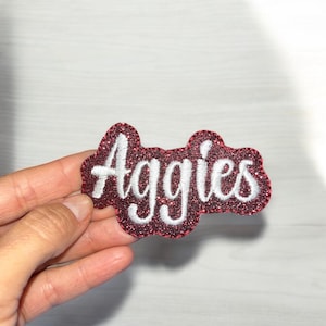 May include: A red glitter patch with the word "Aggies" in white script. The patch has a cloud-like shape and is held by a hand against a white background. The patch is likely for clothing or accessories.