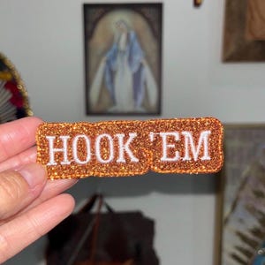 May include: A rectangular orange glitter patch with the words "HOOK 'EM" embroidered in white. The patch is held up in front of a wall with framed artwork.