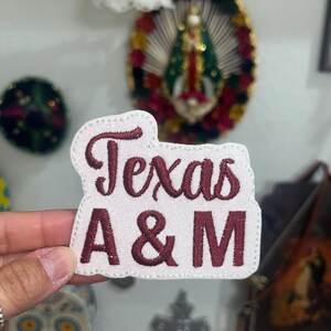 May include: A white embroidered patch with a glittery finish featuring the text Texas A&M in dark maroon thread. The patch has a die-cut shape that follows the contour of the lettering and is finished with a stitched border around the entire edge.