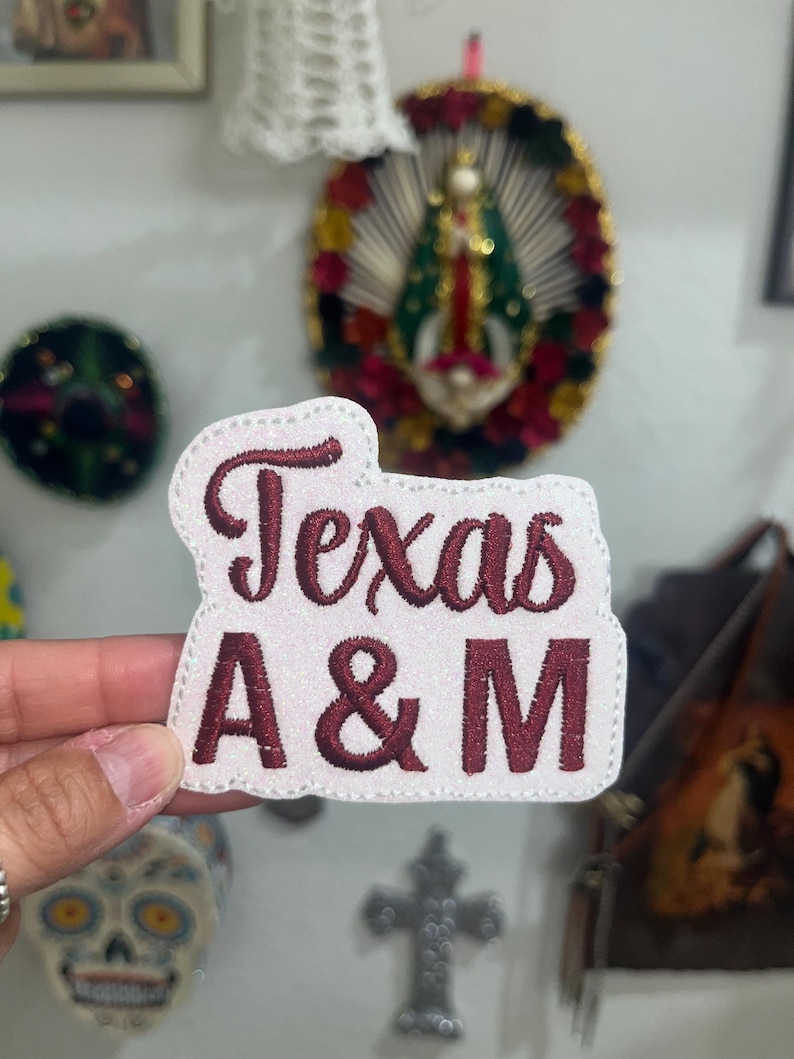 May include: A white embroidered patch with a glittery finish featuring the text Texas A&M in dark maroon thread. The patch has a die-cut shape that follows the contour of the lettering and is finished with a stitched border around the entire edge.