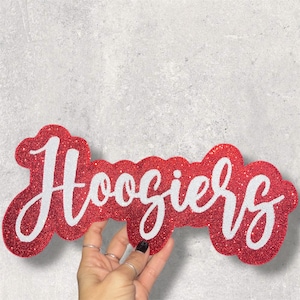 May include: A red glittery sign with the word "Hoosiers" in white script. The sign is held by a hand with rings and a black painted fingernail. The background is a light gray textured surface.