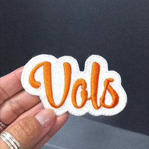 May include: A white patch with orange embroidered text that reads "Vols". The patch has a cloud-like shape with a white border and is held by a hand. The background is a dark gray color.
