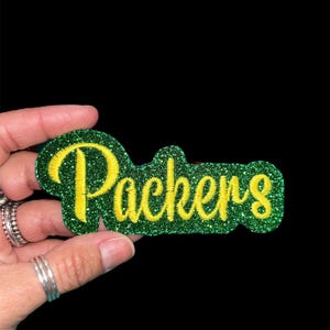 Op de afbeelding: Een groen en gele glitter patch met het woord "Packers" in een script lettertype. De patch is groen met gele letters en een glittertextuur. De achtergrond is zwart.