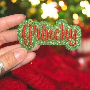 May include: A green glittery applique with the word "Grinchy" embroidered in red. The applique is held in a hand against a blurred background of Christmas lights and red fabric. The word is in a cursive font.