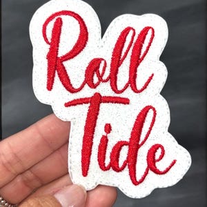 May include: A white patch with red embroidered text that reads "Roll Tide." The patch has a glittery border and is held against a dark background. The text is in a cursive font.