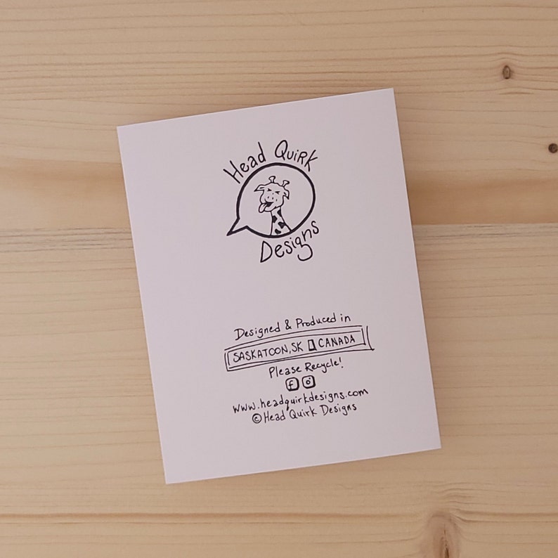 May include: A white card with black text and a cartoon giraffe. The text reads "Head Quirk Designs" and includes information about the product being designed and produced in Saskatoon, SK, Canada. It also encourages recycling and includes the website and copyright information for Head Quirk Designs.