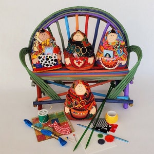 May include: Four colorful fabric pincushions shaped like people are sitting on a whimsical, multi-colored, wooden bench. The pincushions are decorated with various patterns and have pockets for holding sewing supplies. There are also sewing supplies on the table in front of the bench, including needles, thread, and a small wooden spool.