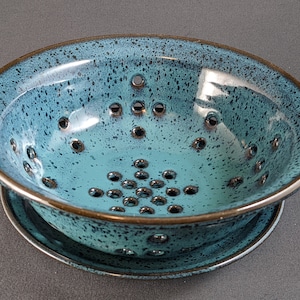 May include: A blue ceramic colander with a matching saucer. The colander has a speckled glaze and a dark brown rim. It has a pattern of round holes in the bottom.