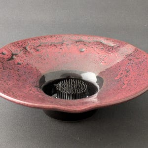 May include: A red ceramic flower frog with a black base. The dish has a textured surface and a circular opening in the center to hold the floral arrangement. The flower frog is a decorative piece for floral design.