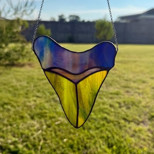 May include: A stained glass suncatcher shaped like a shark tooth, featuring a top section of iridescent blue and purple glass and a bottom section of bright yellow glass, all held together by dark metal solder lines and suspended by a metal chain.