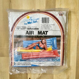 May include: A clear plastic package containing The Wet Set 72 inches by 27 inches inflatable air mat. The packaging features text, a barcode, and a photograph of a person in a blue bikini lying on a blue inflatable mat in a pool.