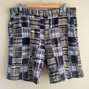 May include: Patchwork shorts featuring a multi-colored plaid pattern in shades of navy blue, olive green, white, and grey. The shorts have a button closure at the waistband, belt loops, and side pockets, constructed from various square fabric panels.