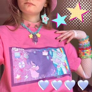 May include: A pink t-shirt with a graphic print of a cartoon character with a retro-futuristic aesthetic. The shirt features the text "thymic cool" and a purple and blue color scheme. The model is wearing a pink choker with silver spikes, a turquoise chain necklace with yellow star charms, and a pair of pastel pink earrings.