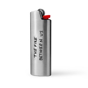 Puede incluir: Un encendedor plateado con el texto "THE FIRE BETWEEN US" grabado en el lateral.