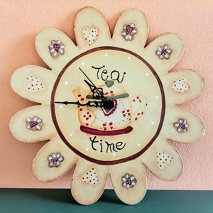 Puede incluir: Un reloj de pared decorativo con forma de flor de 12 pétalos. Cada pétalo tiene un diseño diferente, incluyendo corazones, flores y puntos. El centro del reloj presenta una tetera con las palabras "Tea Time" escritas debajo.
