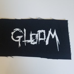 May include: A rectangular black patch with the word "GLOOM" in white, stylized, jagged lettering. The patch has a raw, unfinished edge, suggesting it's ready to be sewn onto fabric. The background is a plain white surface.