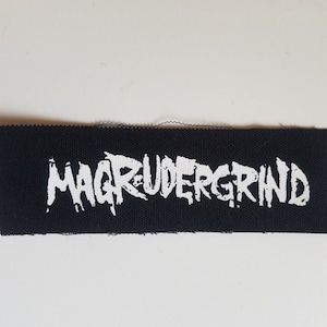 Może przedstawiać: Czarna łatka z tkaniny z białym tekstem, który brzmi "MAGRUDERGRIND".
