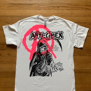 May include: White t-shirt featuring a black and white graphic of a hooded figure holding money. The graphic includes the text "ANTICHRIST" and "I'LL BUY ALL THE URANIUM YOU'VE GOT..."
