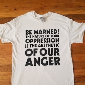 May include: White t-shirt with black text that reads "BE WARNED! THE NATURE OF YOUR OPPRESSION IS THE AESTHETIC OF OUR ANGER"