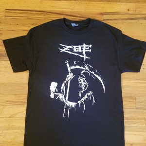 May include: Black t-shirt featuring a white graphic of a skeletal figure holding a scythe and a bird. The graphic also includes the text "XOF".