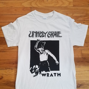 May include: White t-shirt with a black and white graphic print of a person holding a weapon with the text "Unholy Grave" and "Wrath" printed below the image.