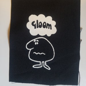 May include: Black square fabric patch with a white cartoon character and the word "gloom" in a cloud-shaped speech bubble. The character has a round body, small feet, and a worried expression.