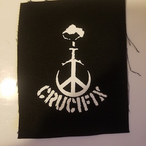 May include: Black fabric patch with a white peace sign with a sword through it and the word "CRUCIFY" written below it.