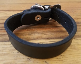 May include: A black leather bracelet with a silver buckle. The bracelet is lying on a wooden surface.