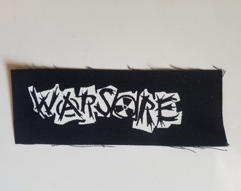 May include: Black rectangular patch featuring the word "WARSORE" in white, jagged lettering. A white radiation symbol is included in the design. The patch has a raw, unfinished edge.