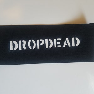 Peut inclure: Écusson en tissu noir avec le texte blanc "DROPDEAD" imprimé dessus.