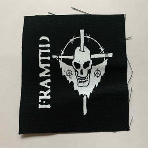 Peut inclure: Écusson carré noir avec un motif blanc de crâne, croix et symboles de paix. Le mot "FRAMTID" est imprimé verticalement sur le côté gauche. L'écusson a un bord brut.