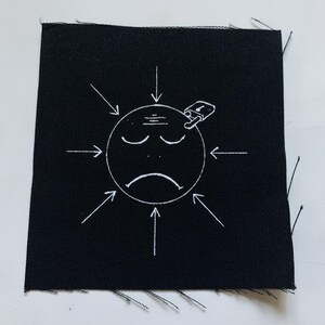 Puede incluir: Parche de tela negro con un contorno blanco de una cara triste con un candado en la cabeza. Las flechas apuntan hacia la cara desde todos los lados.