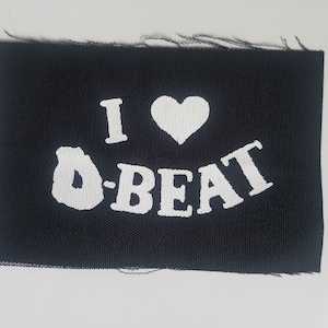 Op de afbeelding: Zwarte stoffen patch met witte tekst die "I ♥ O-BEAT" zegt.