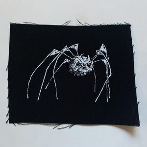 May include: Black fabric patch with a white embroidered image of a spider-like creature with a skull-like face.