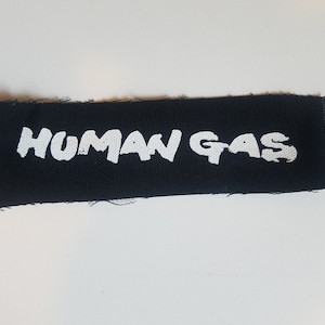 Op de afbeelding: Zwarte stoffen patch met witte tekst die "HUMAN GAS" luidt.