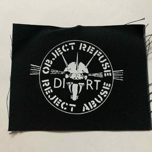 May include: Black fabric patch with white text and a graphic. The text reads "OBJECT REFUSE DIRT REJECT ABUSE" in a circular pattern around a central design. The design appears to be a stylized skull with sharp lines. The edges of the patch are frayed.