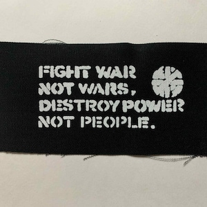 Könnte beinhalten: Schwarzer Stoffaufnäher mit weißem Text: "FIGHT WAR NOT WARS, DESTROY POWER NOT PEOPLE." Eine weiße Grafik befindet sich auf der rechten Seite. Der Aufnäher ist rechteckig mit ausgefransten Kanten.