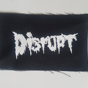 Può includere: Toppa ricamata bianca su tessuto nero con la parola "DISTURBED" in un font stilizzato.