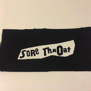 Puede incluir: Parche de tela negro con texto blanco que dice "SORE THROAT" en un estilo irregular dibujado a mano.