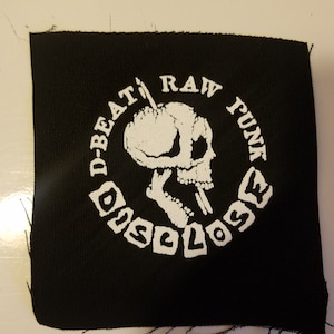 May include: Black fabric patch with white text and a skull graphic. The text reads "D-Beat Raw Punk Disclose".