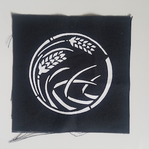Könnte beinhalten: Ein schwarzer Stoff-Patch mit einem weißen Kreisdesign. Der Kreis zeigt ein stilisiertes Weizenähren-Design.