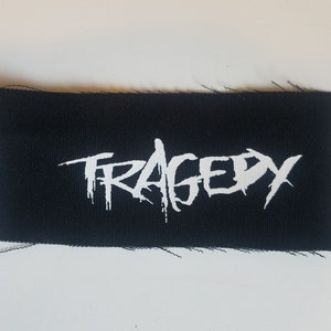 Könnte beinhalten: Schwarzer rechteckiger Aufnäher mit dem Wort "TRAGEDY" in weißer, stilisierter, tropfender Schrift. Der Aufnäher hat einen rauen, unfertigen Rand. Zum Aufnähen auf Kleidung.