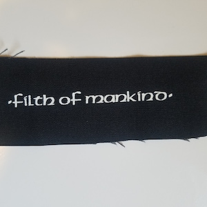 Op de afbeelding: Een zwarte stoffen patch met witte geborduurde tekst die "Filth of mankind." luidt.